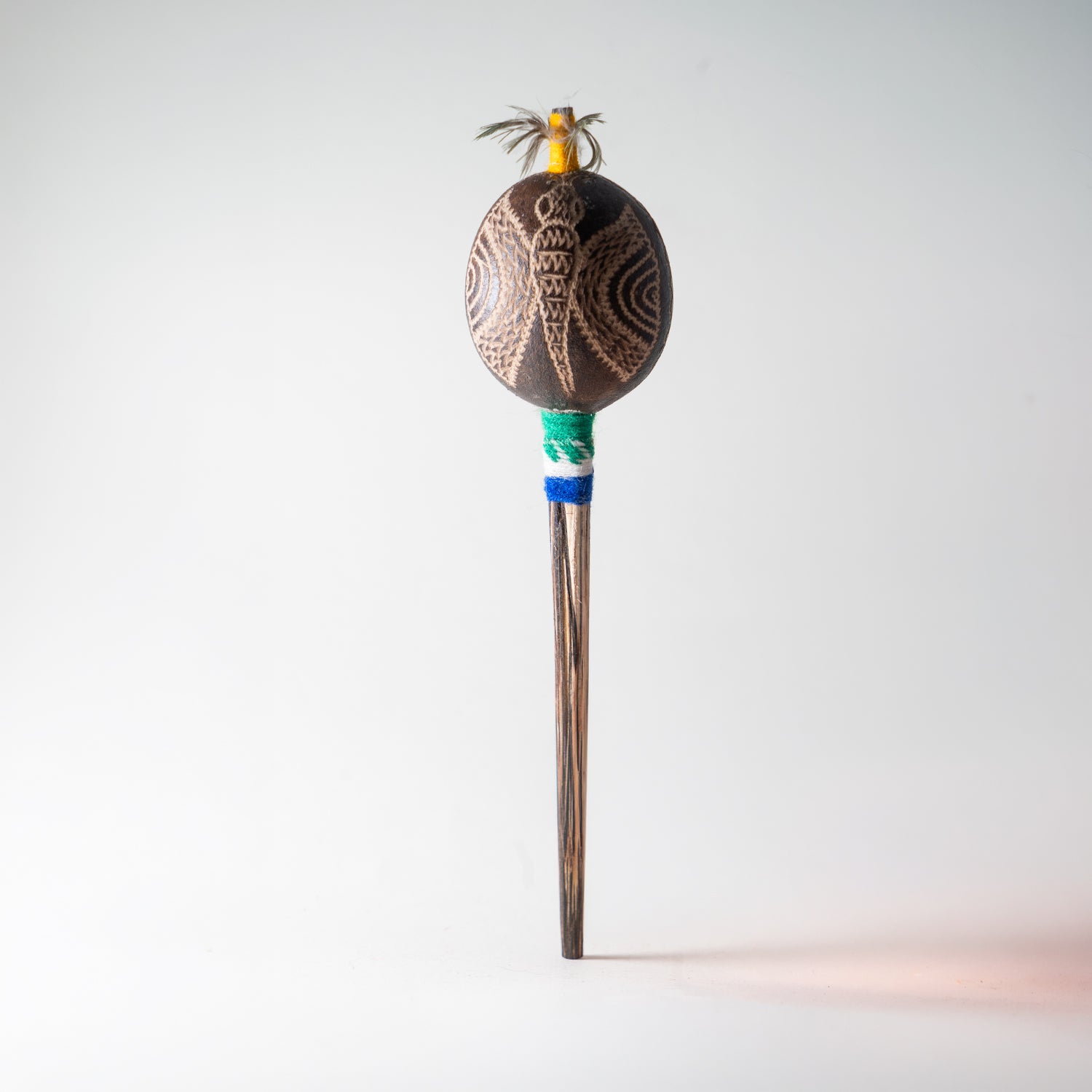 Schamanische Rassel mit eingraviertem Vogelmotiv, gelbem Federschmuck und türkis-blauem Griffband – handgefertigtes Ritualinstrument